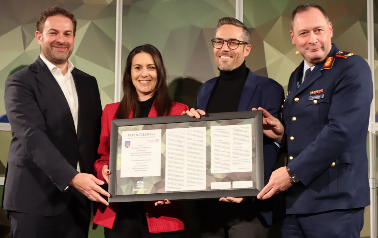 Vier Personen stehen nebeneinander, zwei Männer in Anzug, eine Frau in rotem Blazer und ein Mann in Bundeswehruniform, sie halten gemeinsam einen gerahmten Textdokumentenrahmen