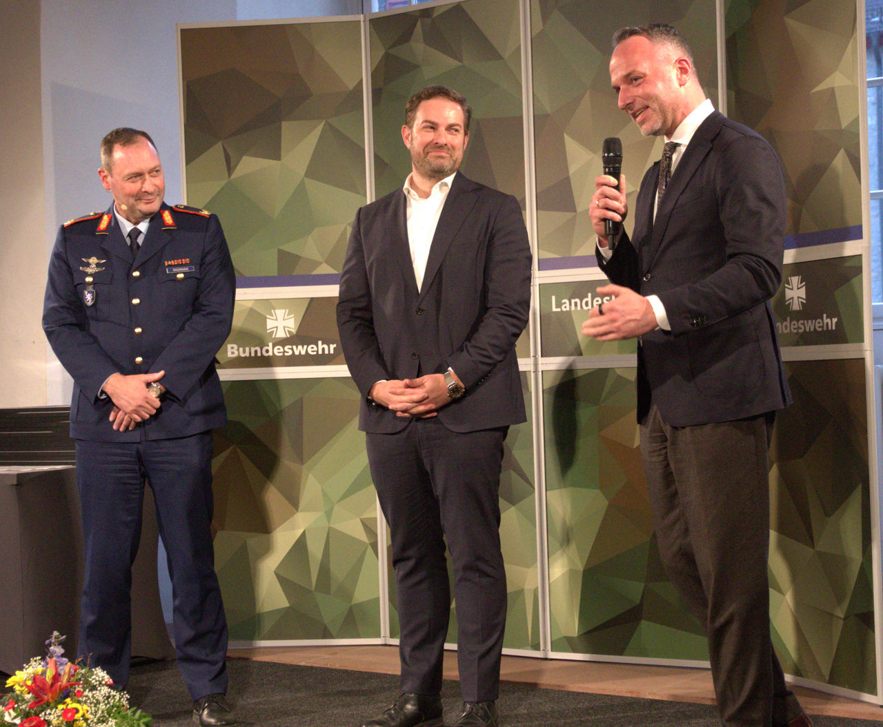 Drei Männer stehen auf einer Bühne vor einer Wand mit Bundeswehr-Logos, einer hält ein Mikrofon und spricht, ein anderer trägt eine Uniform, der dritte einen Anzug.
