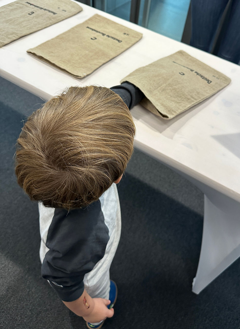 Kind mit blondem Haar steht vor einem Tisch mit drei flachen, rechteckigen, beigen Stoffbeuteln.