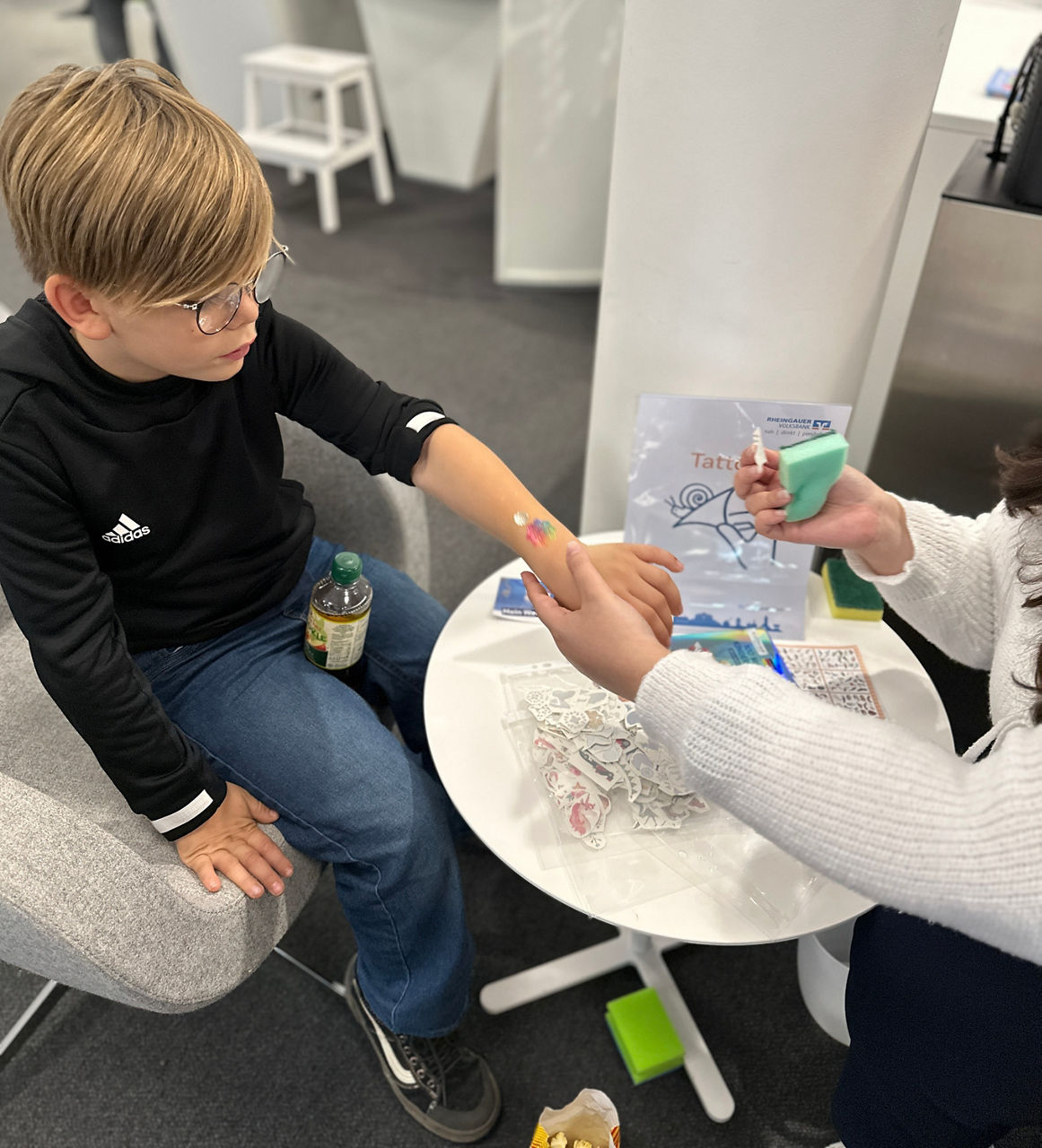 Kind sitzt auf Stuhl, trägt schwarze Jacke und Jeans, bekommt temporäres Tattoo auf Unterarm auf weißem Tisch mit Tattoo-Zubehör aufgestellt.