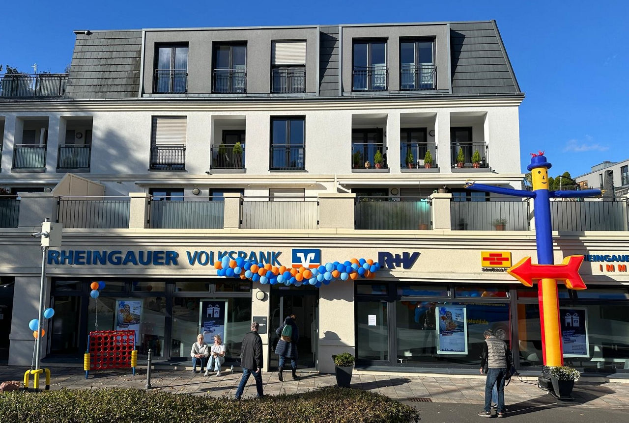 Fassade eines mehrstöckigen Gebäudes mit Balkonen, darunter eine Filiale der Rheingauer Volksbank mit blauen und orangen Luftballons und mehreren Passanten davor.