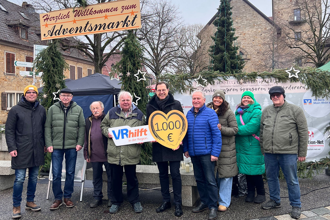 Auf dem Bild stehen 9 Personen nebeneinander auf dem Adventsmarkt. Die Person in der Mitte ist stellvertretender Vorstandsvorsitzender der VR-Bank Main-Rhön, er hält ein Schild in Herzform  mit der Spendenaufschrift darauf. 