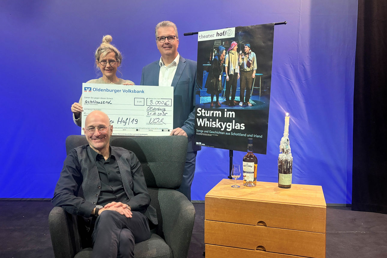 Drei Personen mit einem Bankscheck und einem Plakat vom Theaterstück