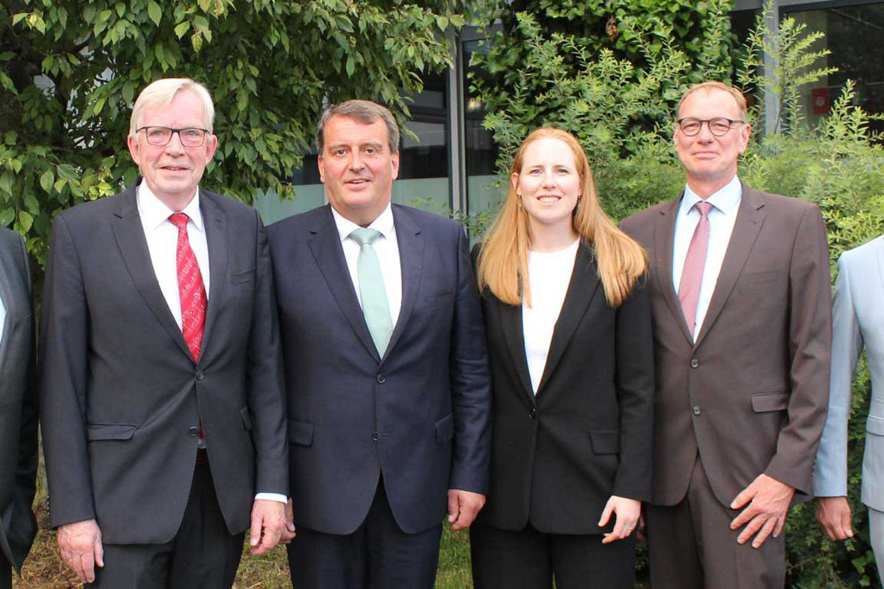 Eine sechsköpfige Gruppe, bestehend aus fünf Männern und einer Frau in Business-Kleidung, posiert lächelnd im Freien vor Büsc