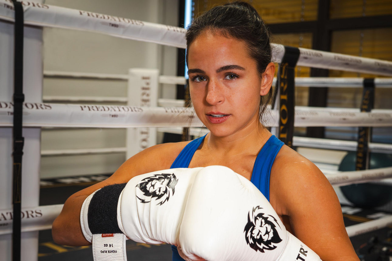 Eine selbstbewusste Boxerin steht mit erhobenen Handschuhen im Boxring und blickt fokussiert in die Kamera.