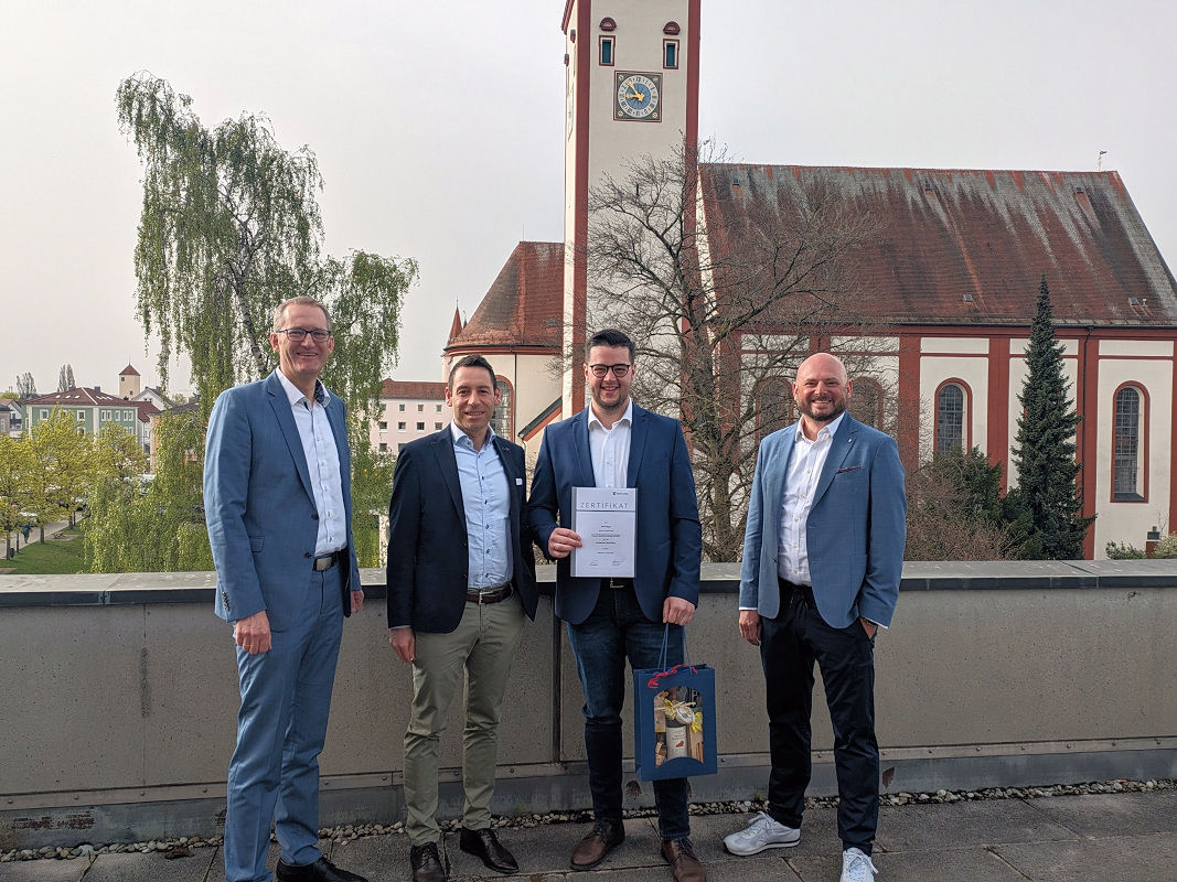 Mitarbeiter steht mit Urkunde und Geschenkkorb neben drei Führungskräften auf einer Dachterrasse mit Kirche im Hintergrund