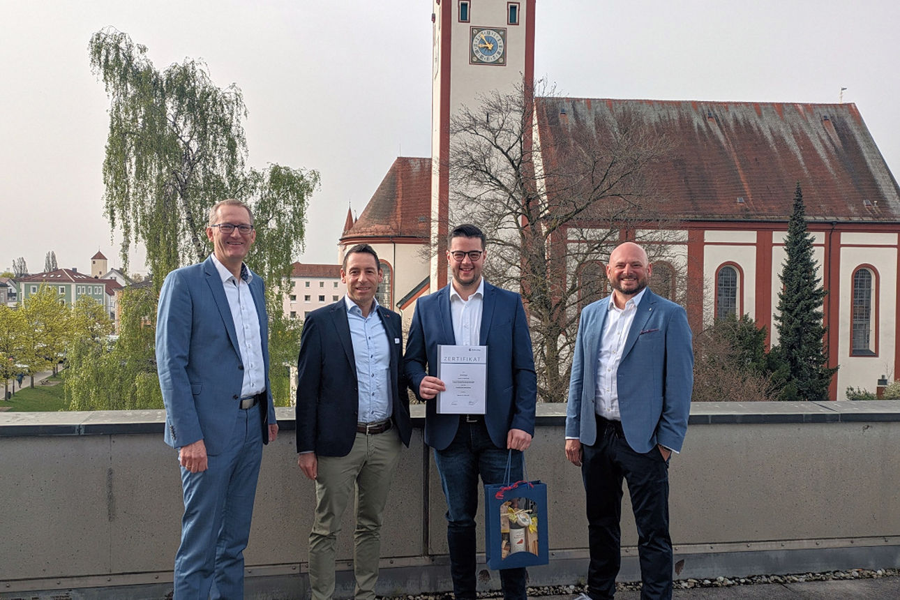 Mitarbeiter steht mit Urkunde und Geschenkkorb neben drei Führungskräften auf einer Dachterrasse mit Kirche im Hintergrund