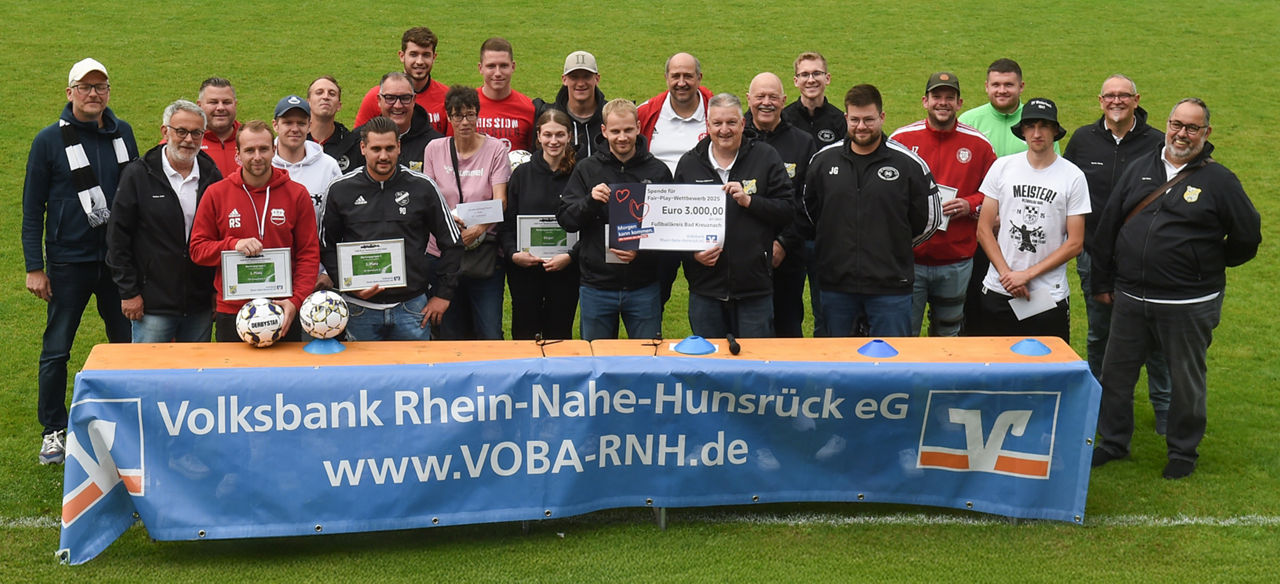 Viele Personen stehen auf einer grünen Wiese. Im Vordergrund steht ein langer Tisch mit einem Banner auf der Vorderseite.