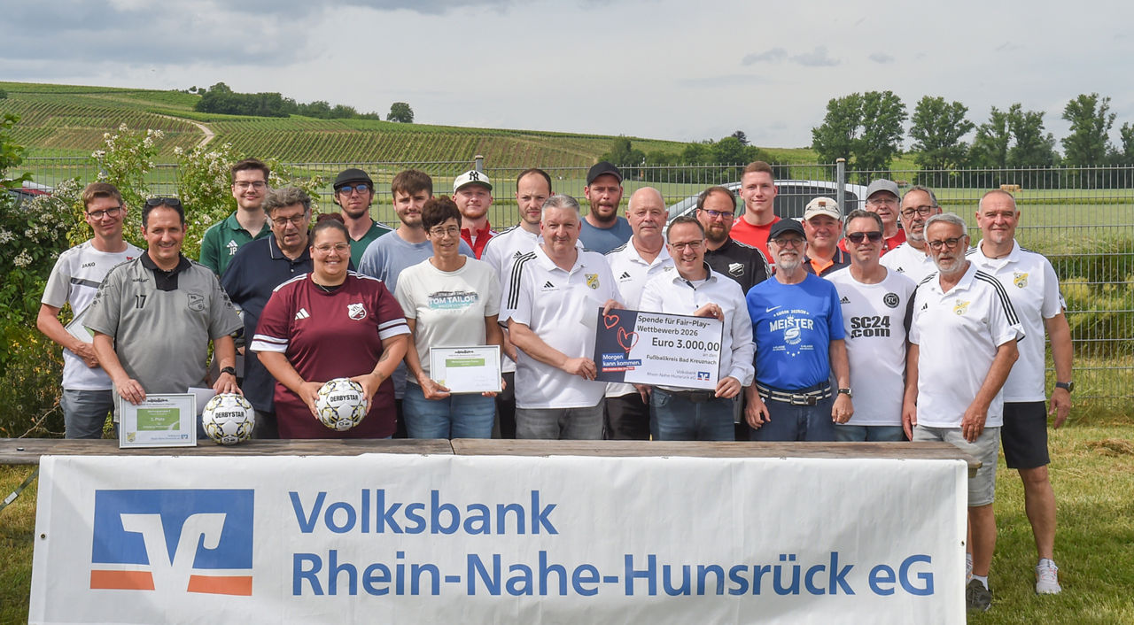 Viele Personen stehen auf einer grünen Wiese. Im Vordergrund steht ein langer Tisch mit einem Banner auf der Vorderseite.