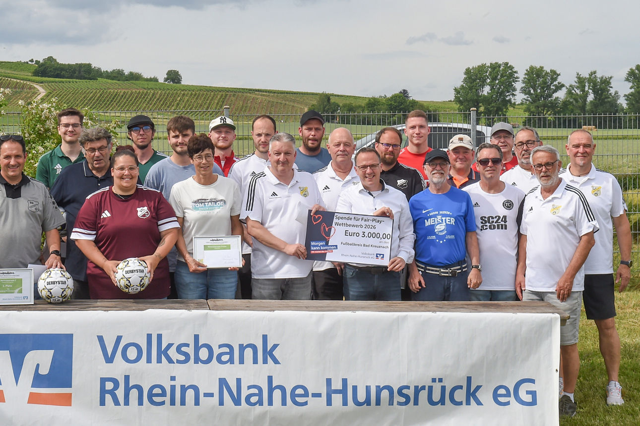 Viele Personen stehen auf einer grünen Wiese. Im Vordergrund steht ein langer Tisch mit einem Banner auf der Vorderseite.
