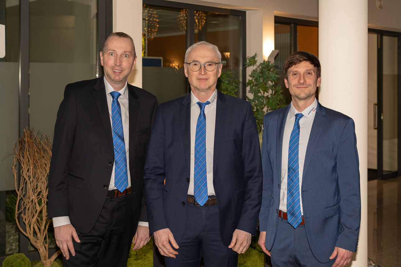 Drei Männer in Business-Anzügen stehen in einer lichtdurchfluteten Umgebung zusammen.