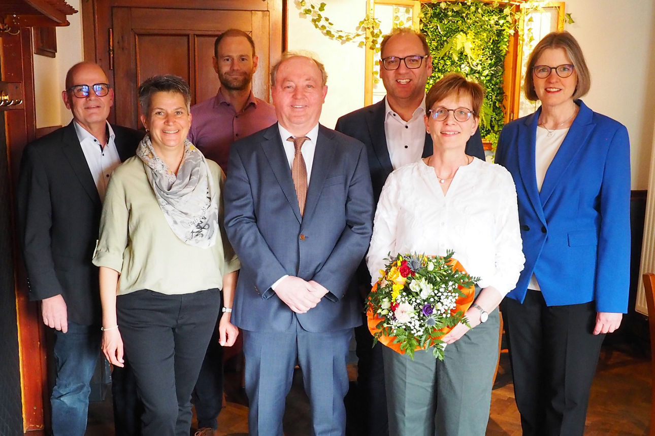 Sieben Erwachsene posieren lächelnd in einem Innenraum; vorne hält eine Frau in weißer Bluse einen bunten Blumenstrauß.