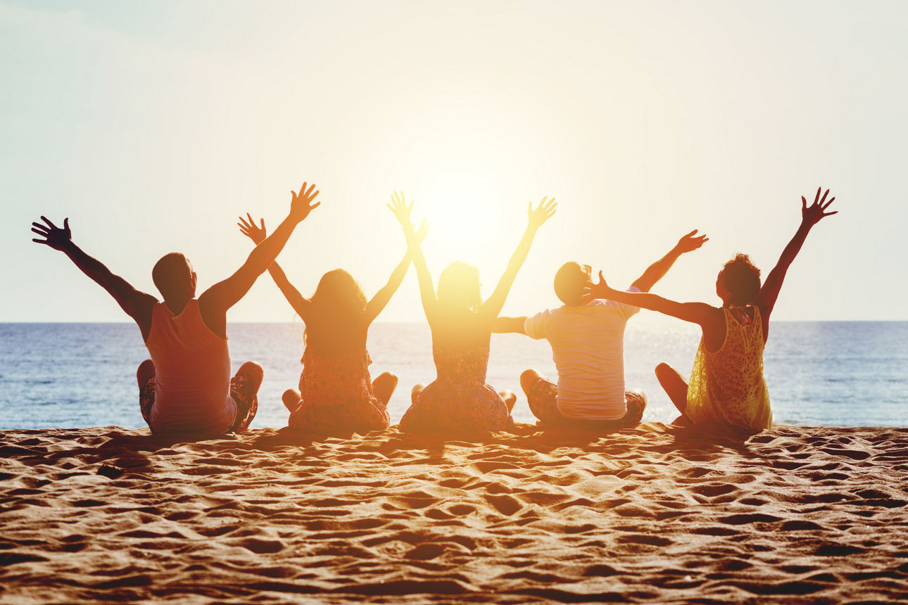 vier junge Menschen sitzen am Strand und heben die Arme in die höhe.