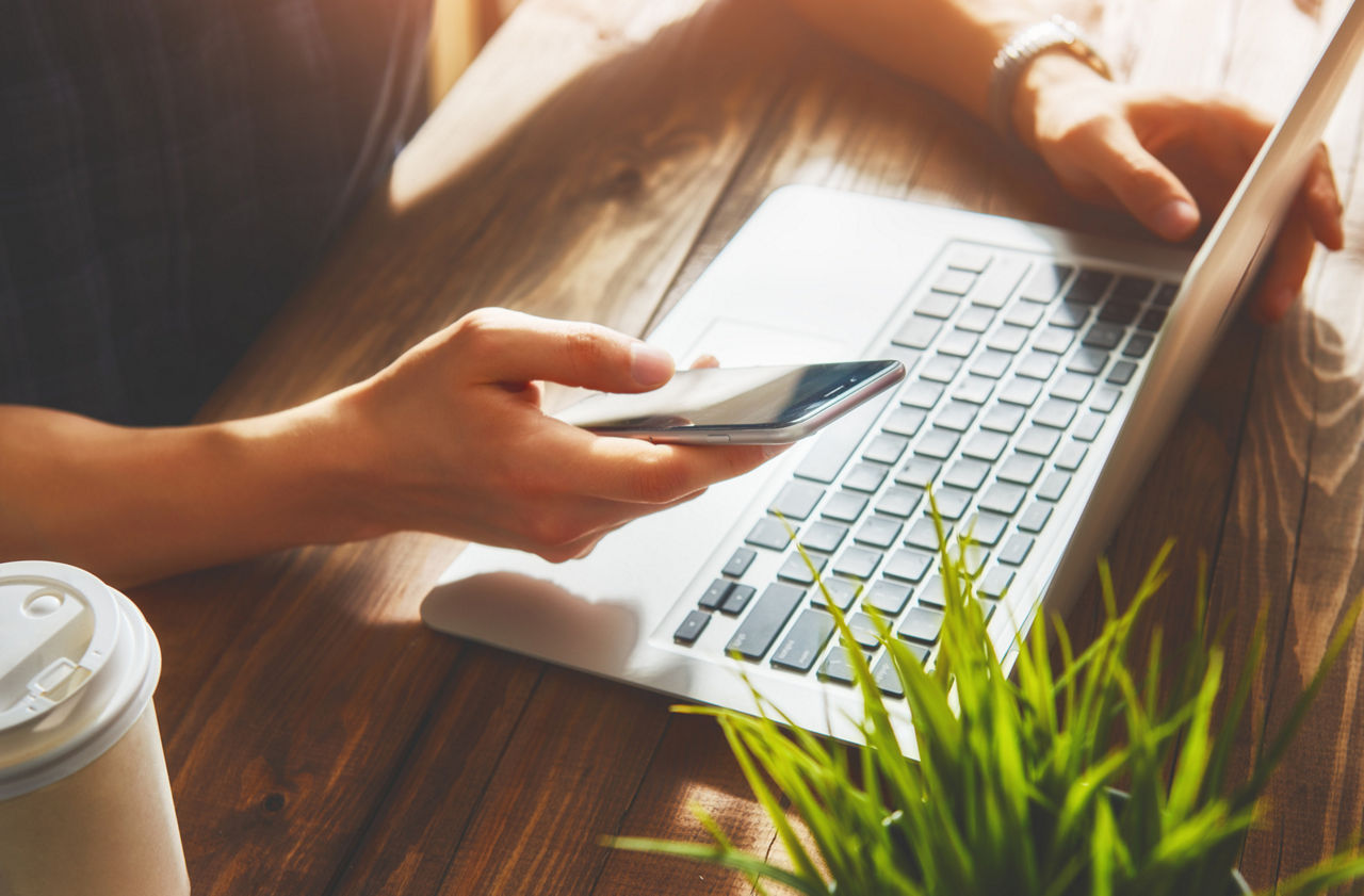 Person am Laptop mit Handy in der Hand