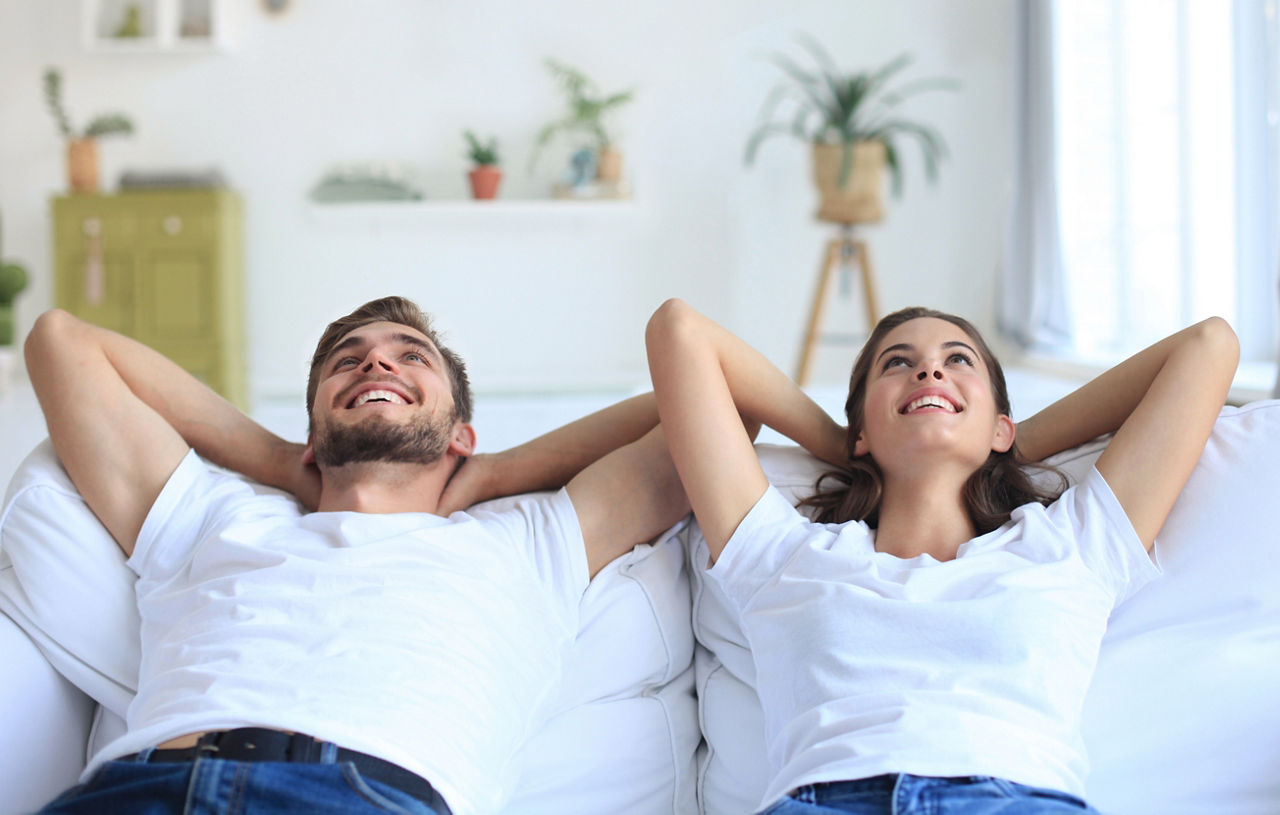 Junges, grlückliches Paar liegt auf dem Sofa, die Arme hinter dem Kopf verschränkt und blickt nach oben
