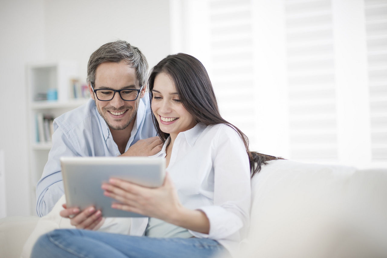 Zwei Personen sitzen auf einem Sofa und schauen gemeinsam auf ein Tablet; im Hintergrund ein Bücherregal und Fensterjalousien