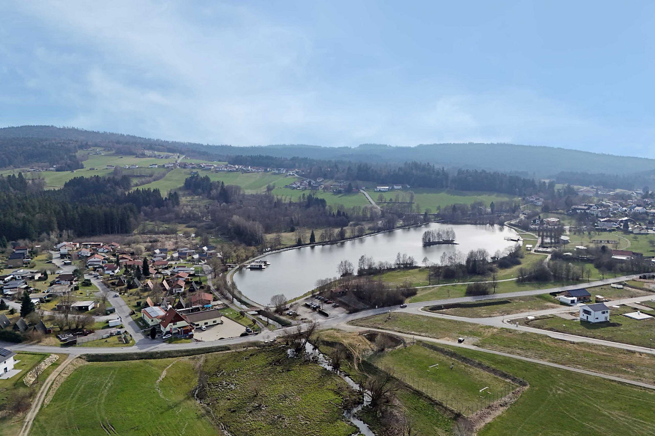 Luftaufnahme einer idyllischen Landschaft mit einem See, umgeben von Häusern, Feldern und Waldflächen unter einem weiten, hel