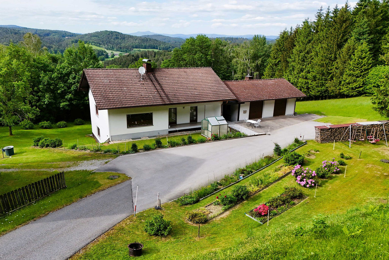 Luftaufnahme eines Einfamilienhauses mit Garage, umgeben von einem grünen Garten, Blumenbeeten und Waldrand in einer hügelige