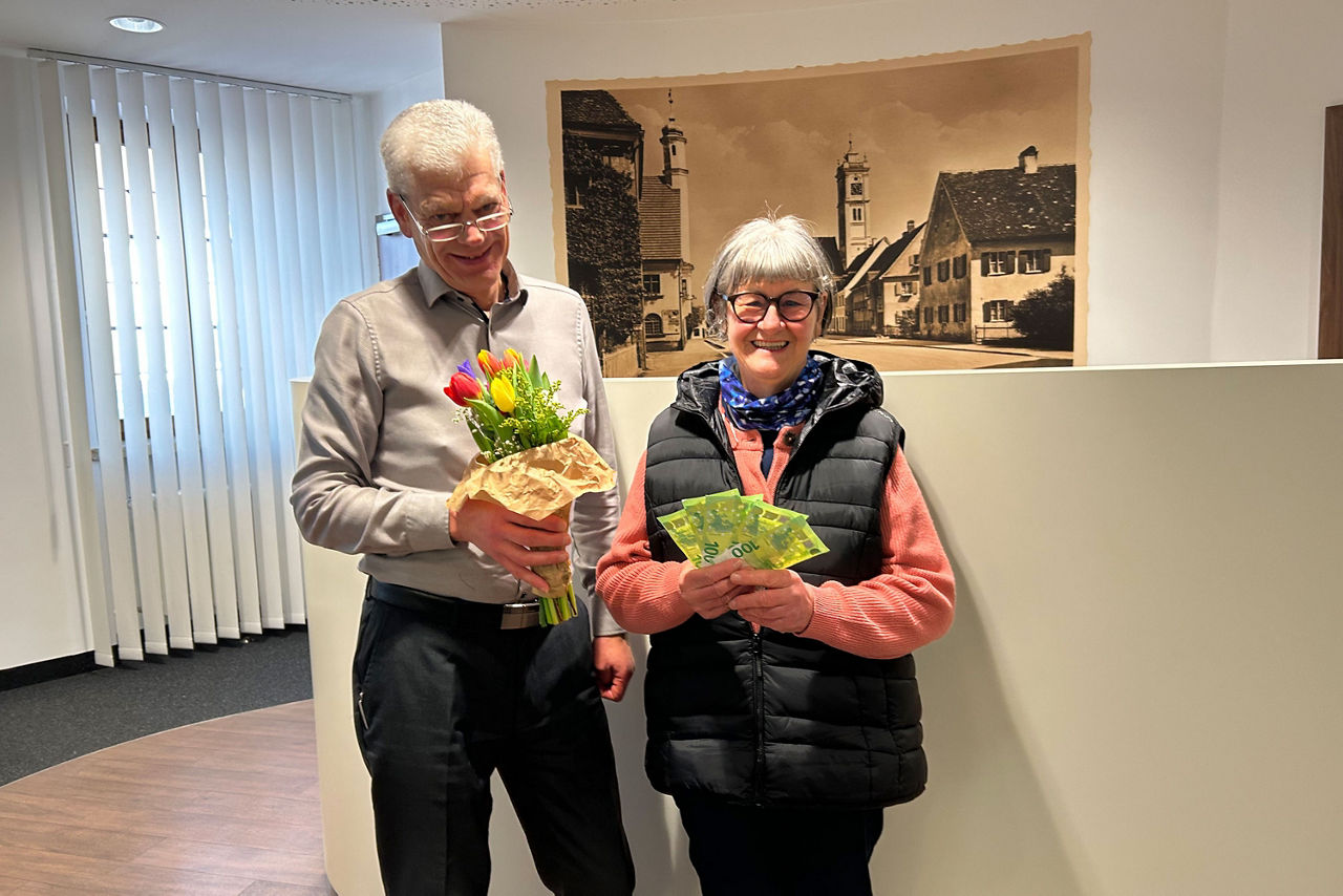 Ein Mann überreicht einer lächelnden Frau einen bunten Tulpenstrauß. Sie hält grüne Scheine in der Hand. Im Hintergrund hängt