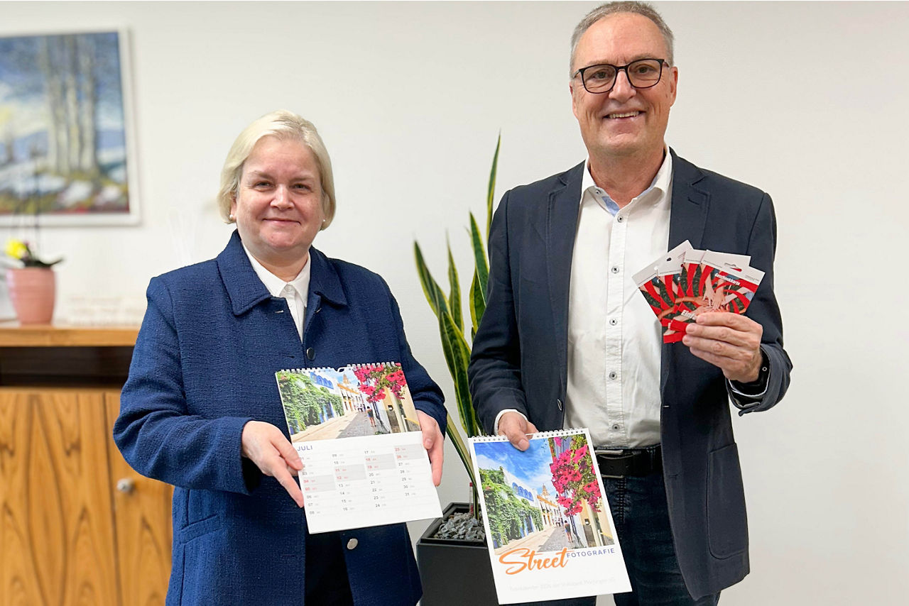  Zwei Personen halten Kalender und mehrere rote Karten, im Hintergrund Pflanze und Gemälde an der Wand.