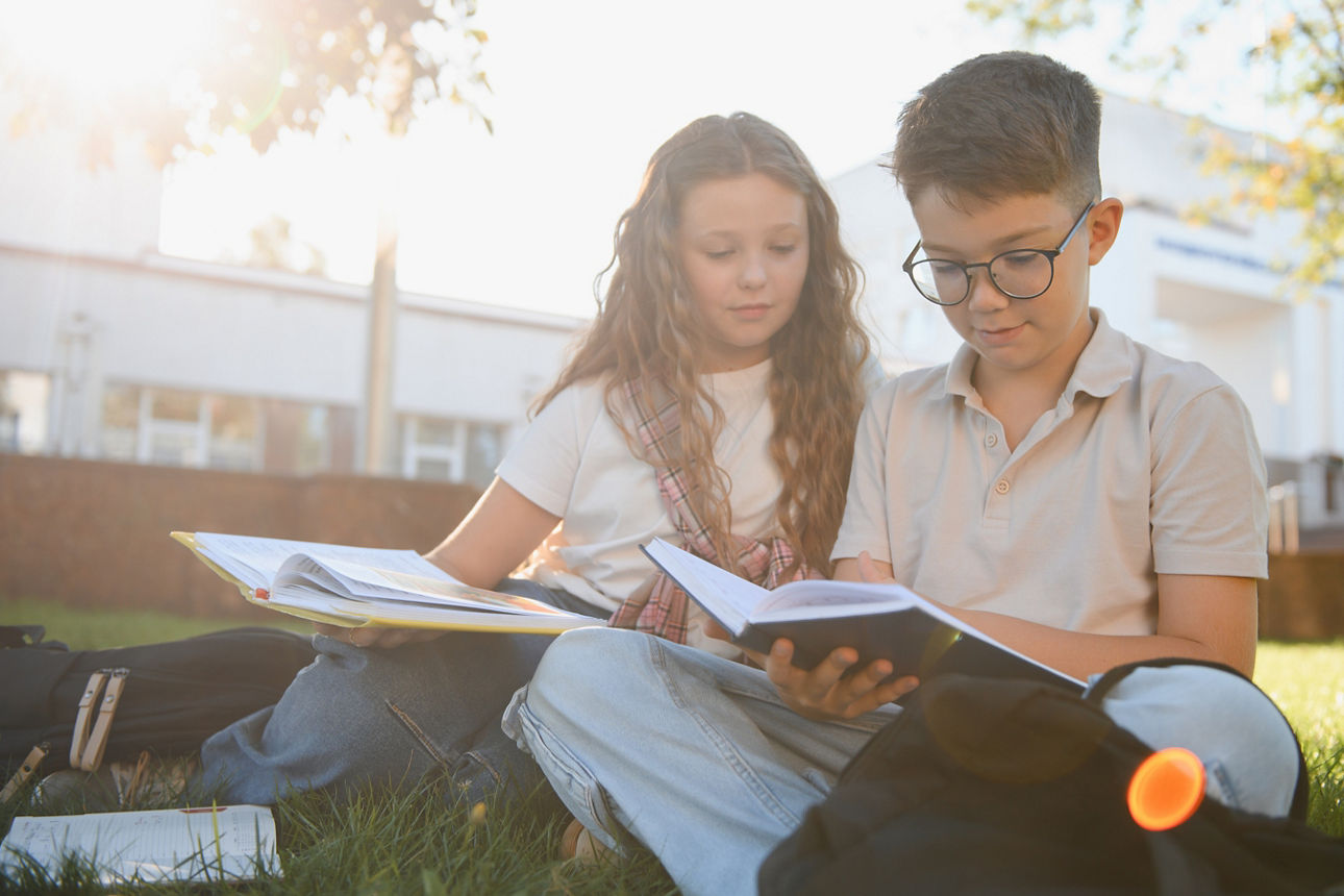Zwei Schulkinder sitzen auf der Wiese vor dem Schulgebäude und lesen in ihren Büchern.