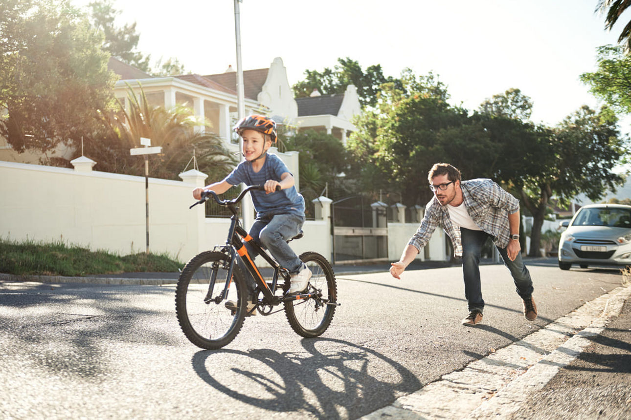 Ein Kind sitzt auf einem Fahrrad und wird von seinem Vater bei der Anfahrt unterstützt