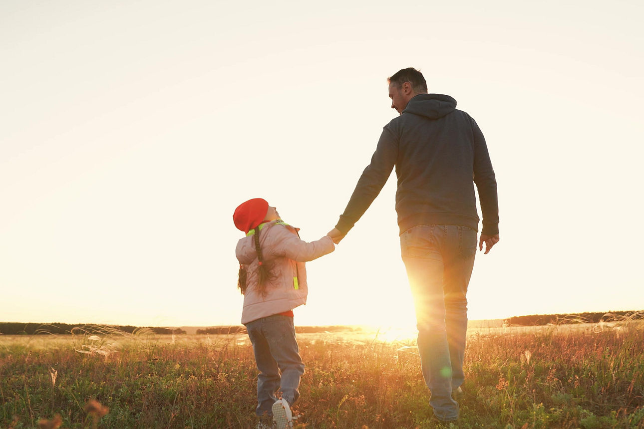 Ein Erwachsener und ein Kind gehen Hand in Hand über eine Wiese bei Sonnenuntergang.