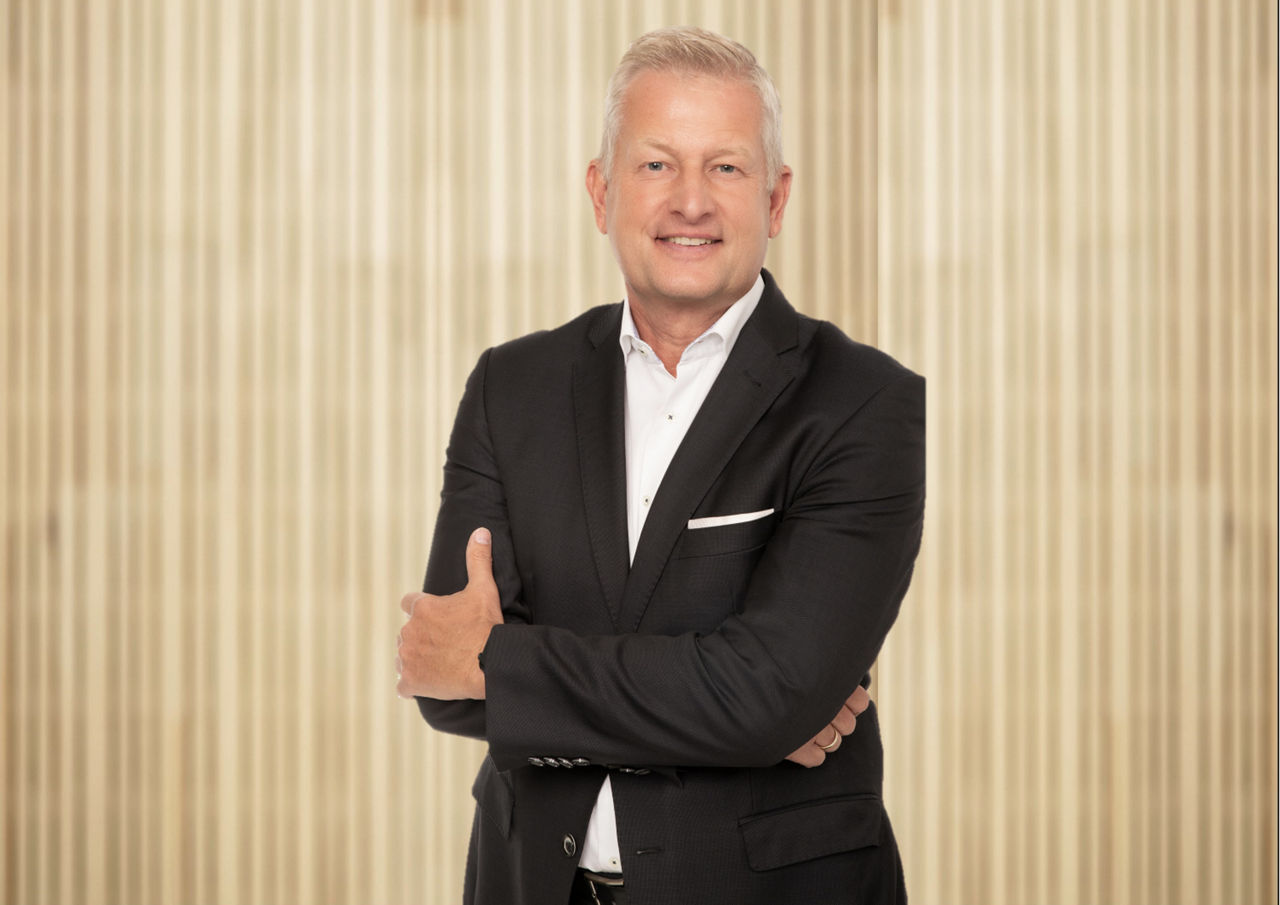 Mann mit schwarzem Sacko steht schräg vor einer Holzwand und verschränkt die Arme