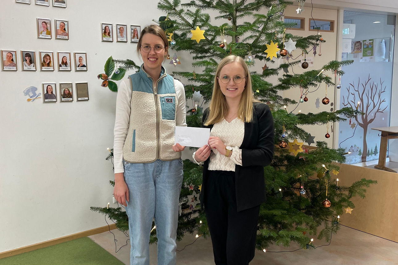 Zwei Frauen lächeln mit einem Briefumschlag in der Hand in die Kamera. Sie stehen vor einem geschmückten Christbaum.