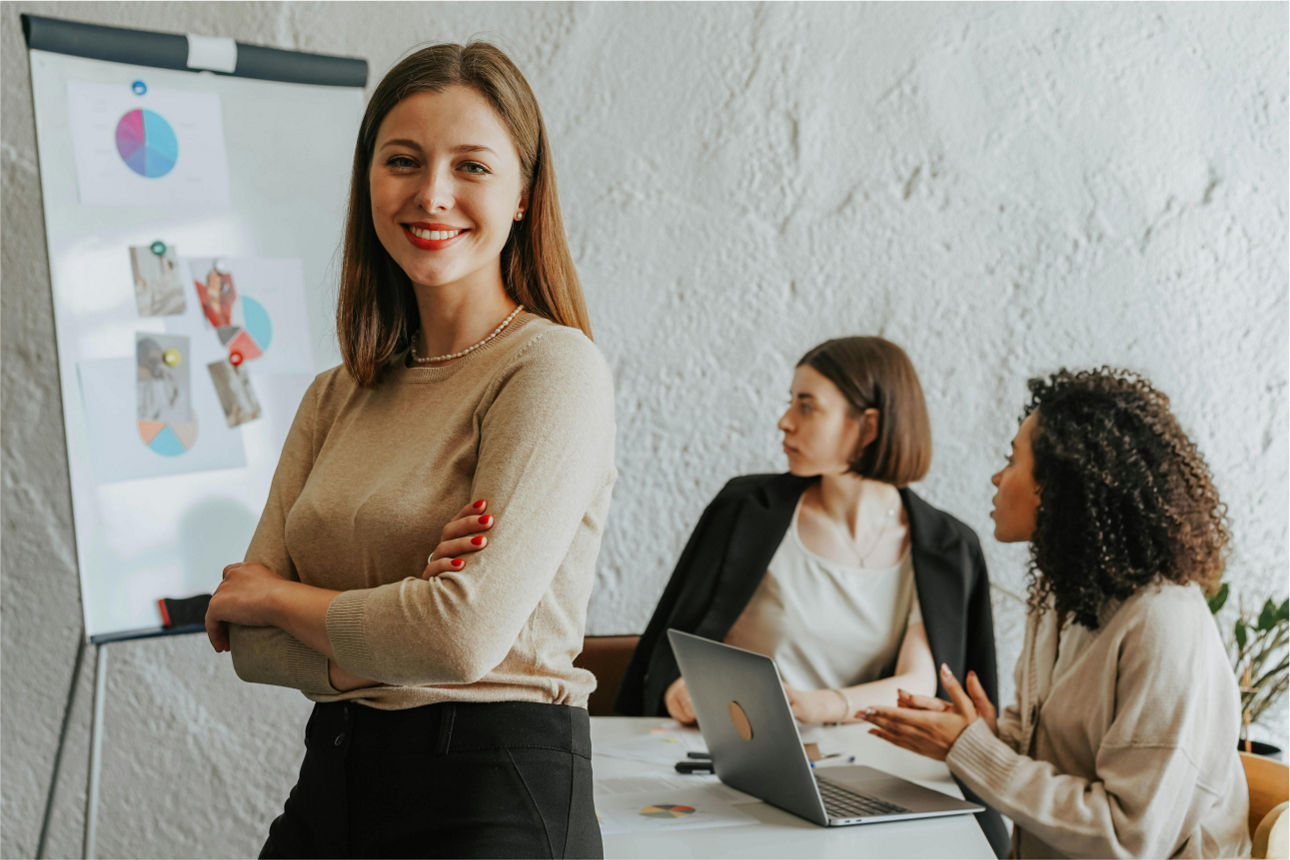 Eine Frau steht vor einem FlipChart und lächelt, im Hintergrund sitzen zwei Frauen die gespannt zuschauen