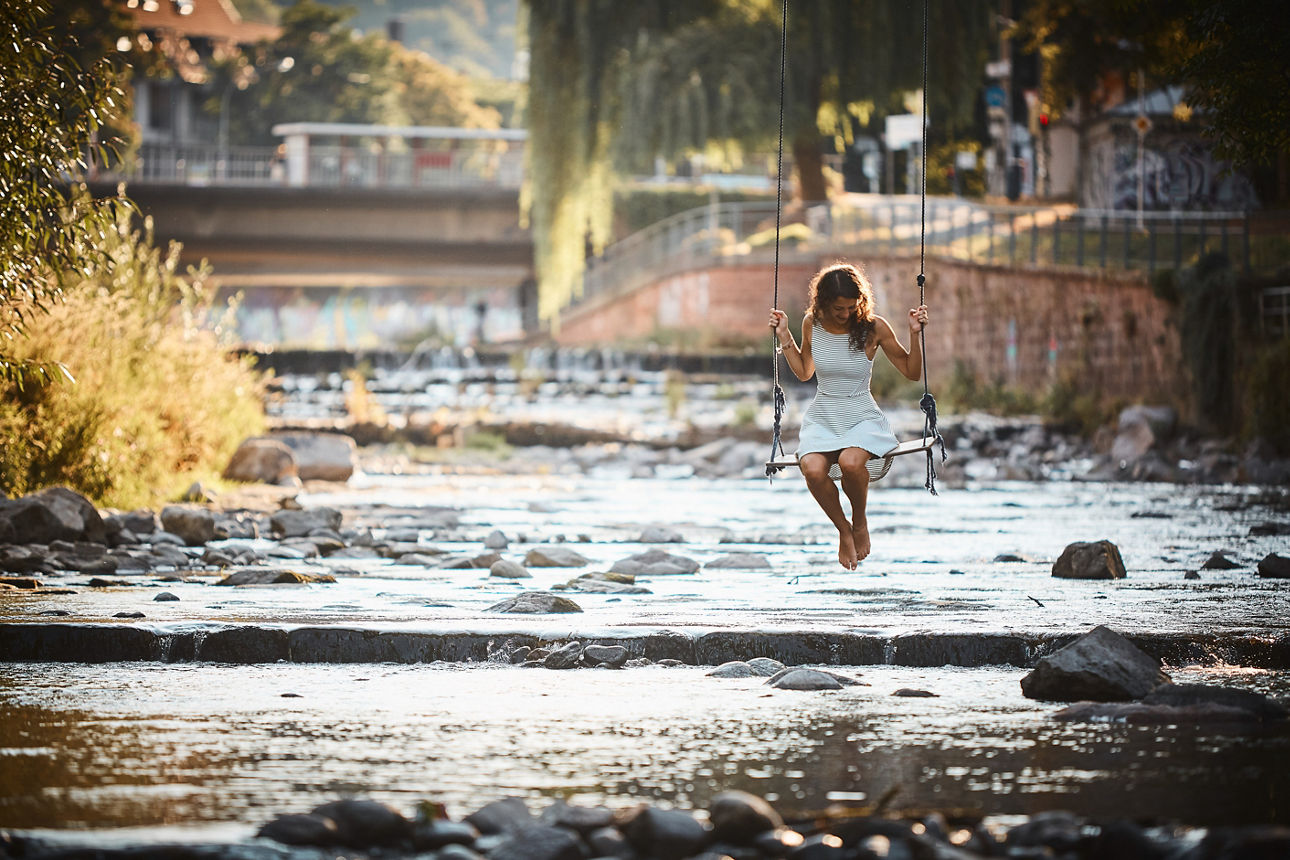 Eine Frau sitzt barfuß auf einer Schaukel über einem flachen Fluss mit Steinen in urbaner Umgebung.