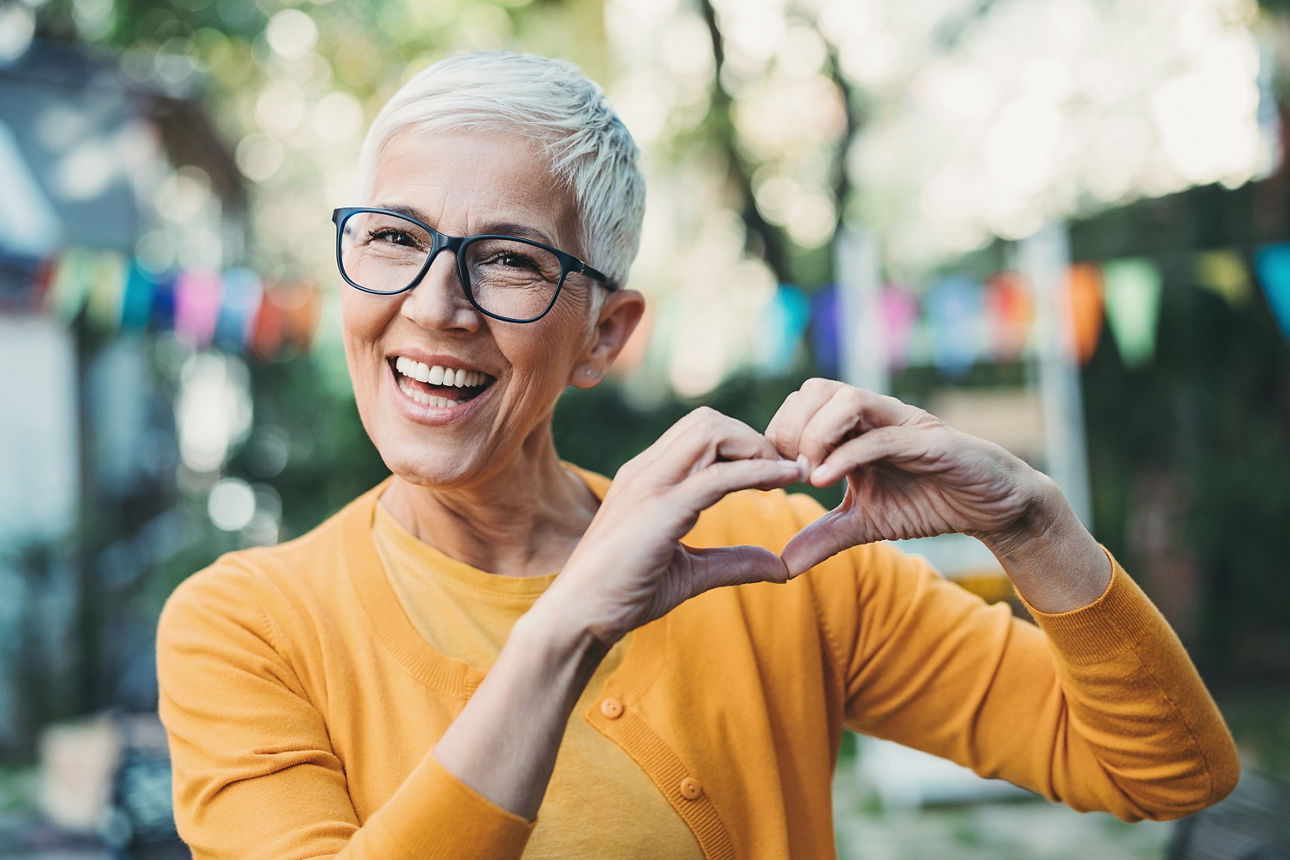 Frau mit kurzen weißen Haaren und einem orangem Oberteil macht ein Herz mit ihren Händen und lacht