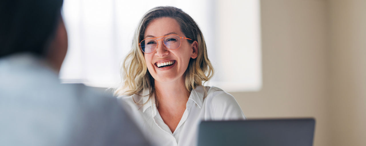 Eine Frau mit orangefarbener Brille sitzt an einem Schreibtisch in einem hellen Raum und spricht mit einer weiteren Person im Vordergrund.