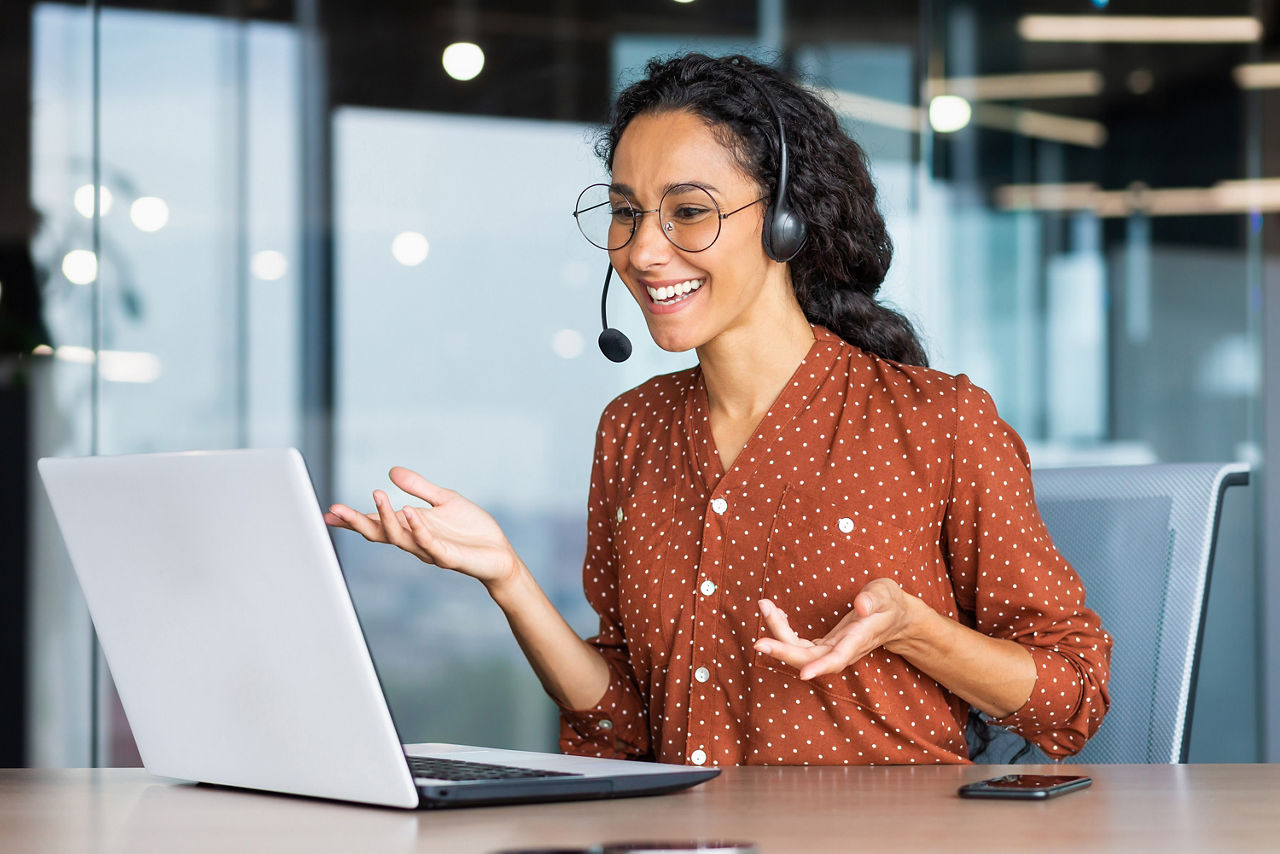 Eine Frau mit dunklen Haare führt ein Telefongespräch über ein Headset an einem Laptop