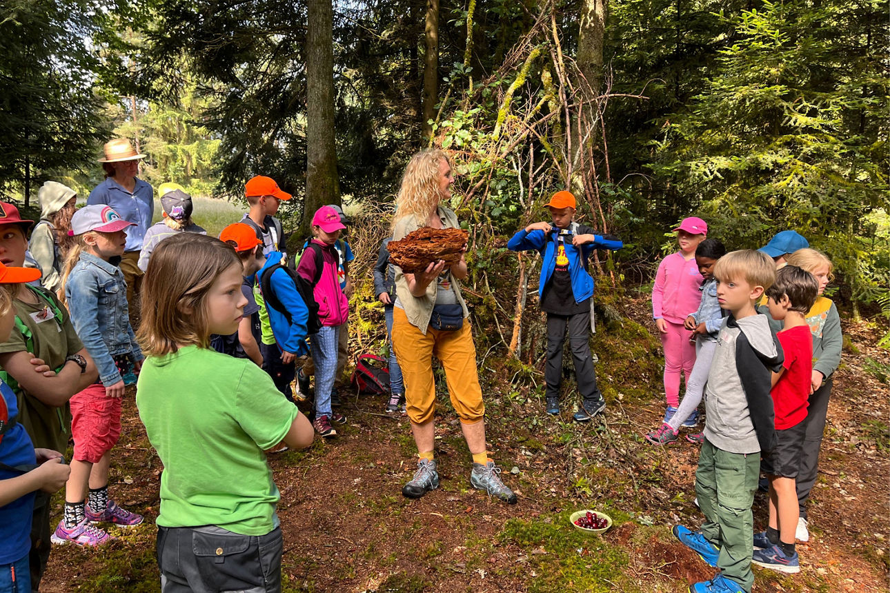 Eine Frau zeigt einer Gruppe von Kindern im Wald ein großes Stück Rinde; sie stehen vor einer selbstgebauten Hütte aus Ästen.