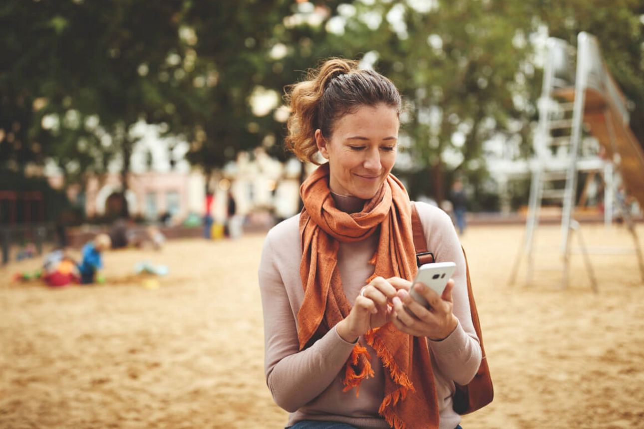 Frau tippt auf ihrem Smartphone auf einem Spielplatz.