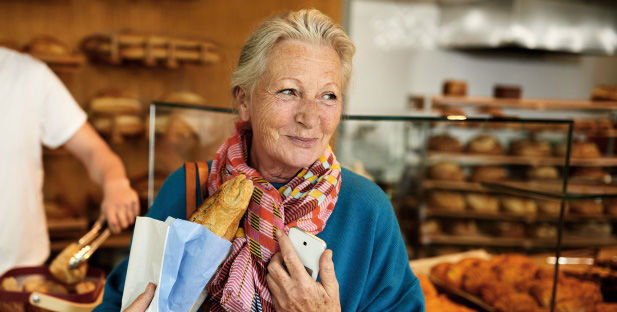Ältere Dame steht beim Bäcker mit einem Baguette und einem Handy in der Hand