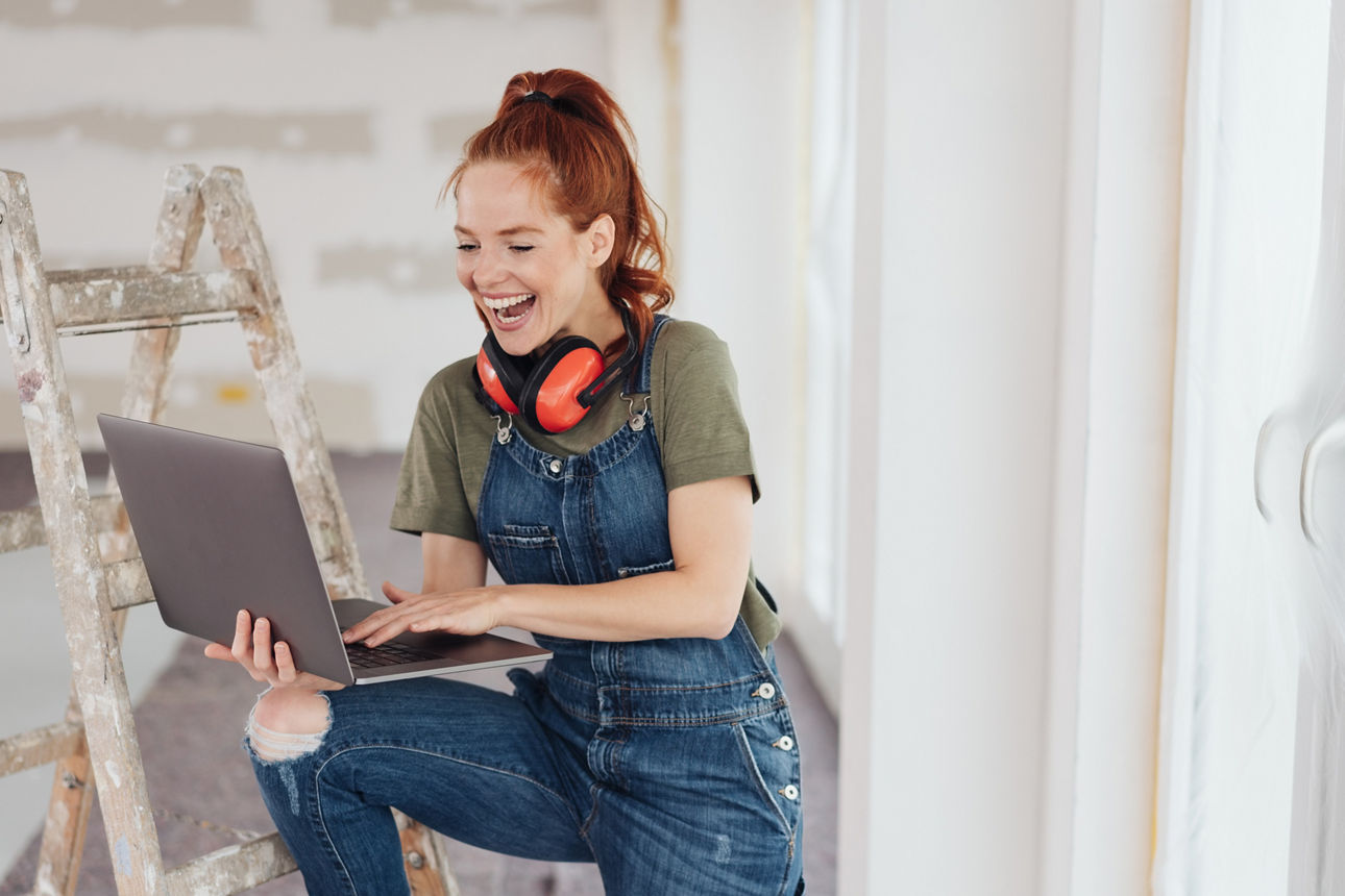 Lächelnde rothaarige Frau mit Laptop in der Hand in Latzhose beim Renovieren