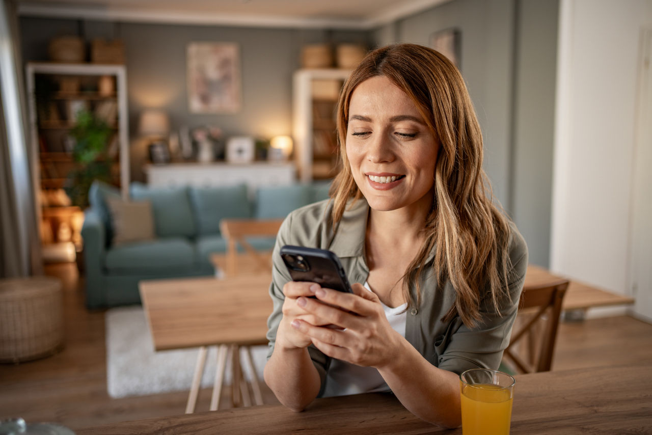 Lächelnde Frau nutzt ihr Smartphone an einem Tisch in gemütlicher Wohnumgebung.