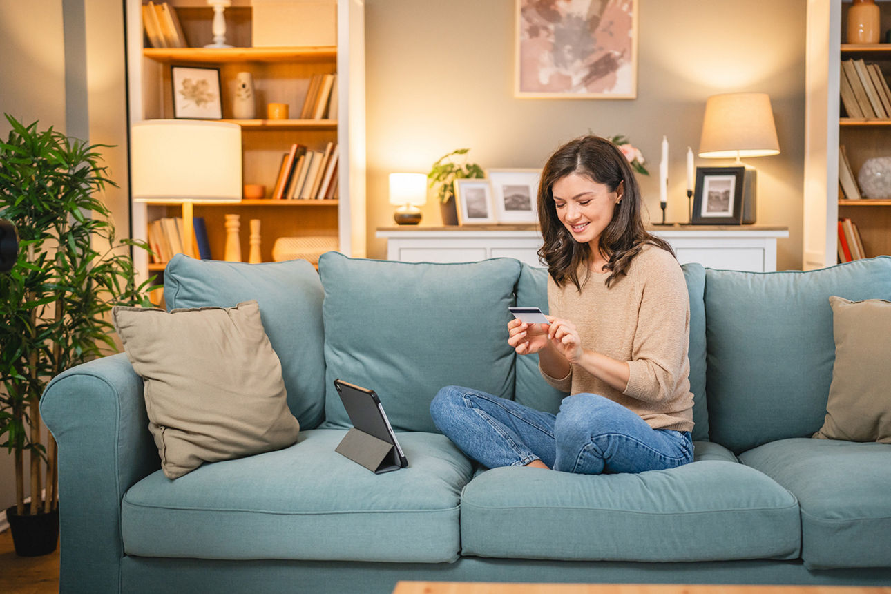 Frau sitzt lächelnd auf einem Sofa, hält eine Bankkarte in der Hand, vor ihr ein Tablet