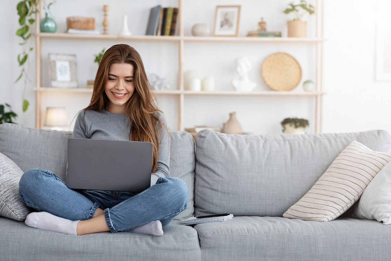 Lächelnde junge Frau, die von zu Hause aus arbeitet, auf der Couch sitzt und ihren Laptop benutzt.