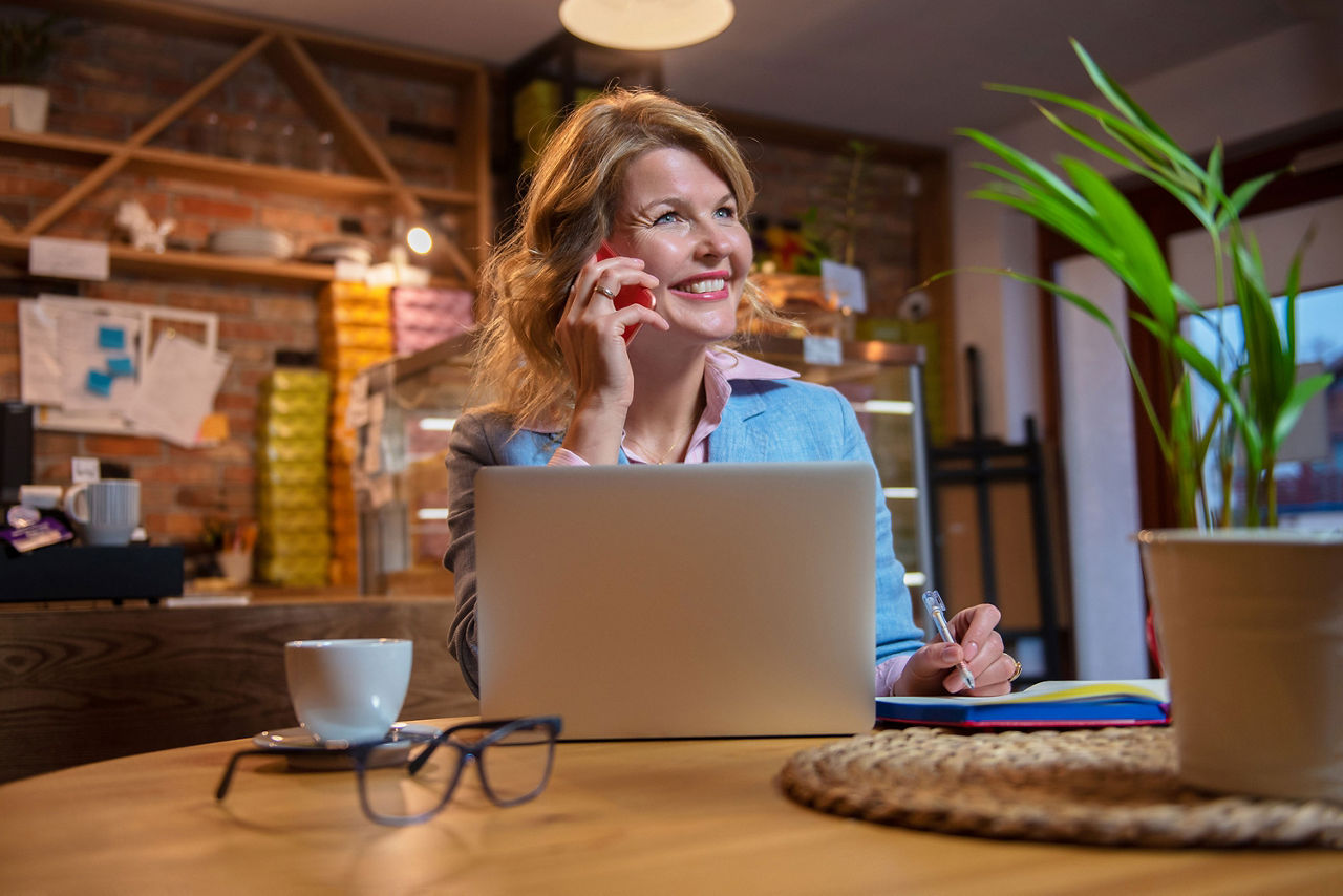 Frau mit lockigem, rotem Haar, lächelnd und mit Smartphone in der Hand, stehend in einer Stadtumgebung.