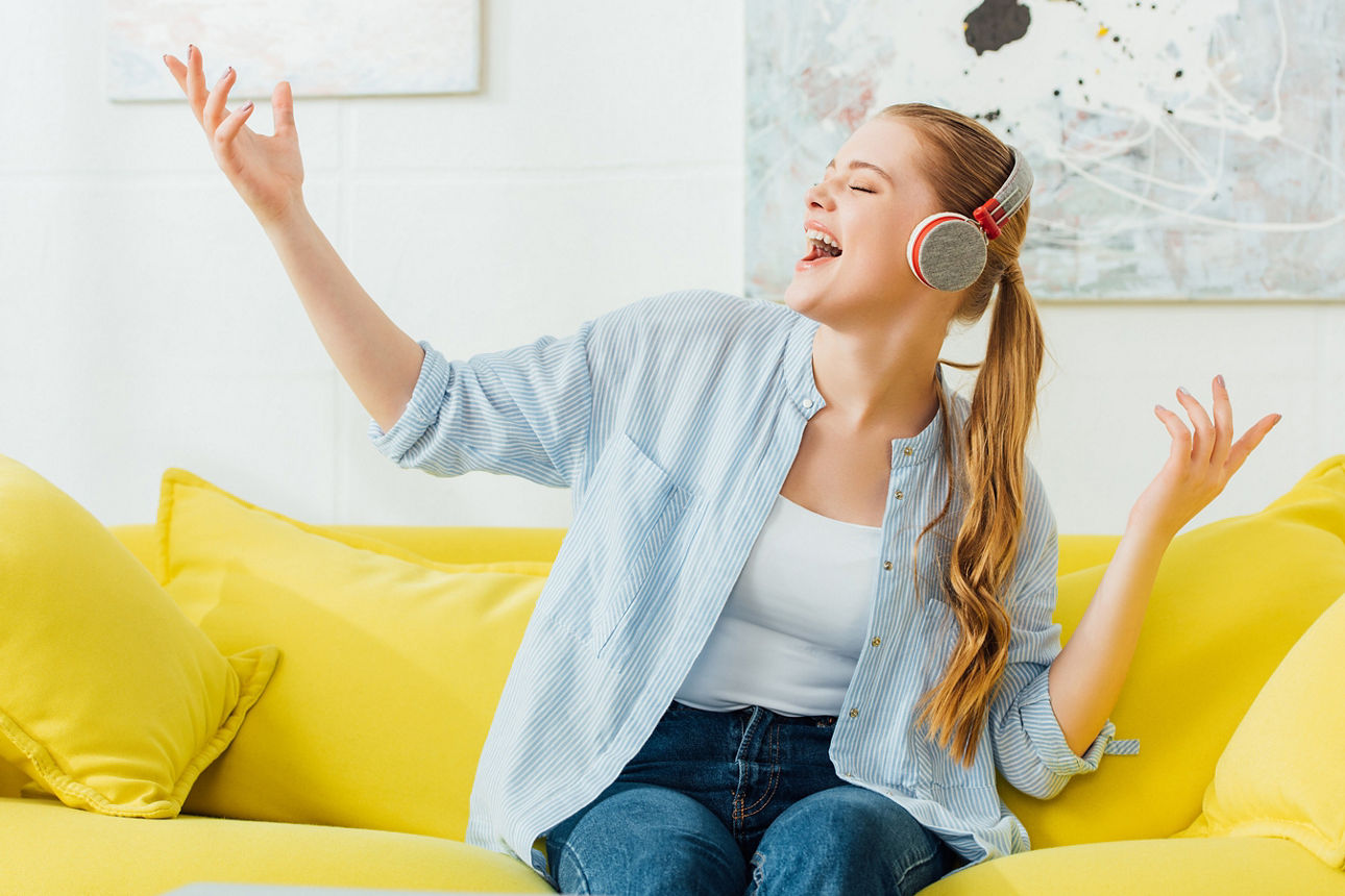 Junge Frau mit Kopfhörern, die auf einer gelben Couch singt, fröhliche Stimmung bei Musik.