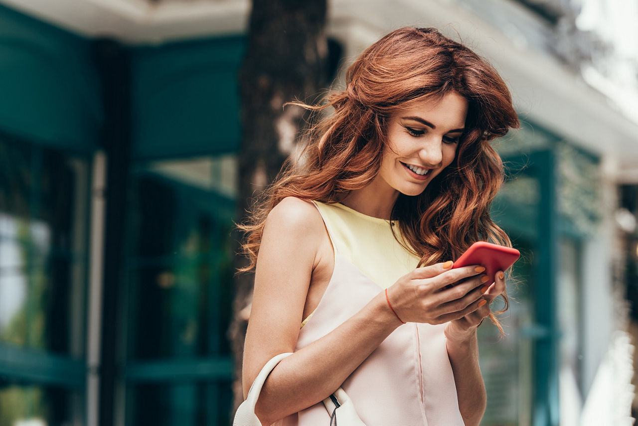 Frau mit lockigem, rotem Haar, lächelnd und mit Smartphone in der Hand, stehend in einer Stadtumgebung.