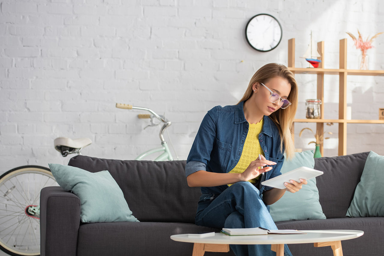 Junge Frau nutzt digitales Tablet, während sie zu Hause auf der Couch in der Nähe des Couchtisches sitzt