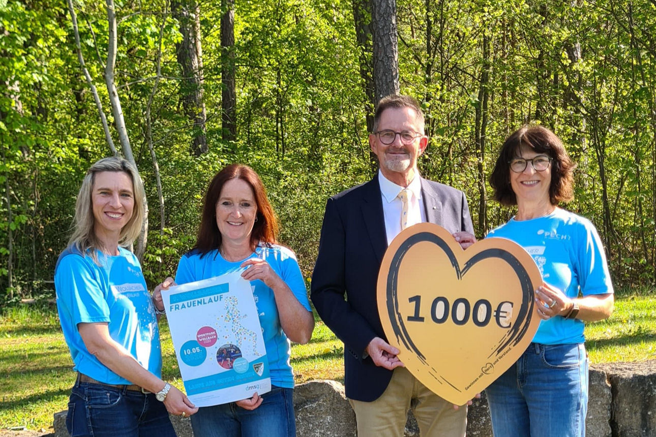 Im Bild stehen drei Frauen und ein Mann im Anzug in einem Waldgebiet. Sie halten ein Schild mit der Aufschrift 1000 Euro.