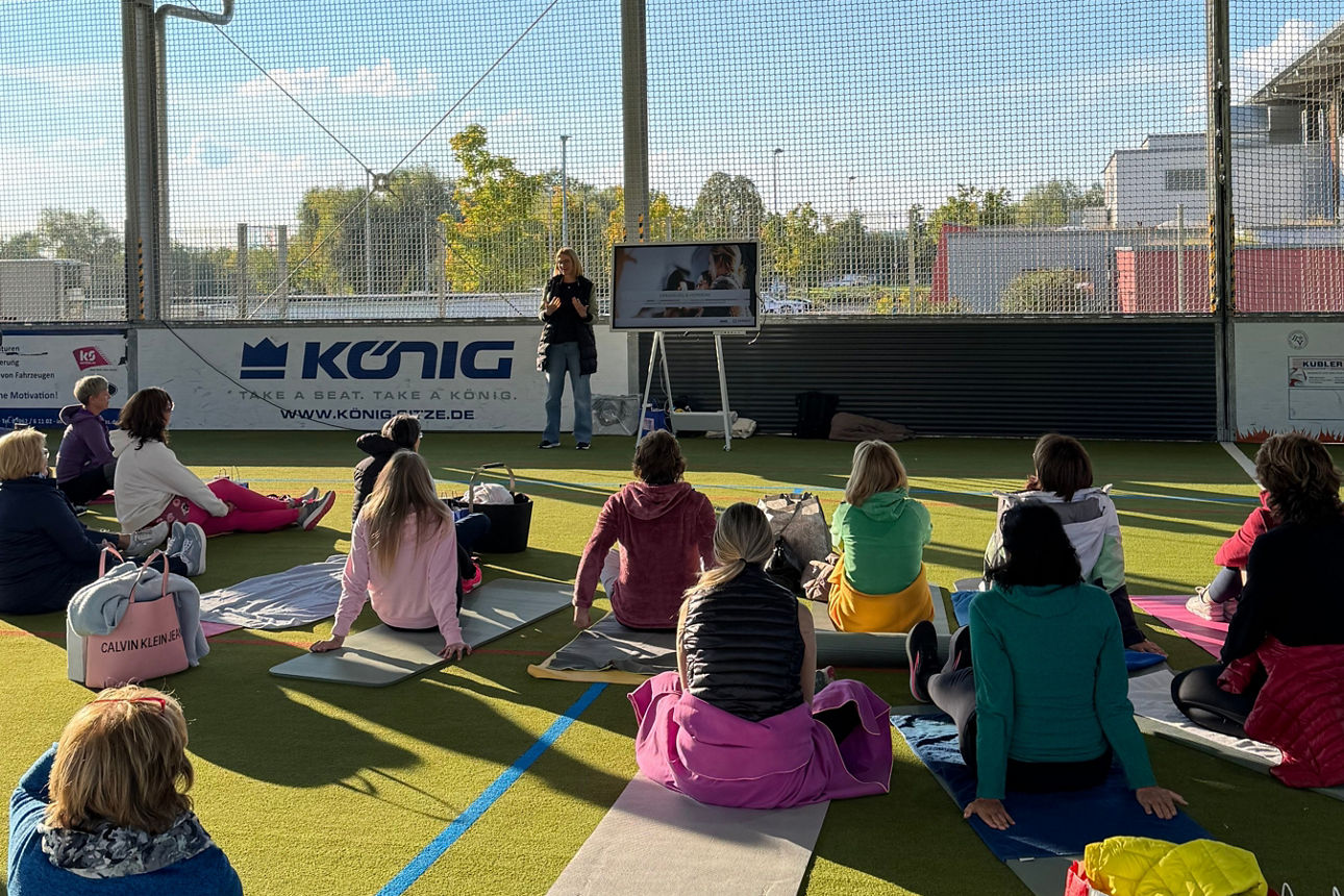Personen sitzen auf Yogamatten auf einem überdachten Sportfeld und schauen einer Präsentation auf einem Bildschirm zu.