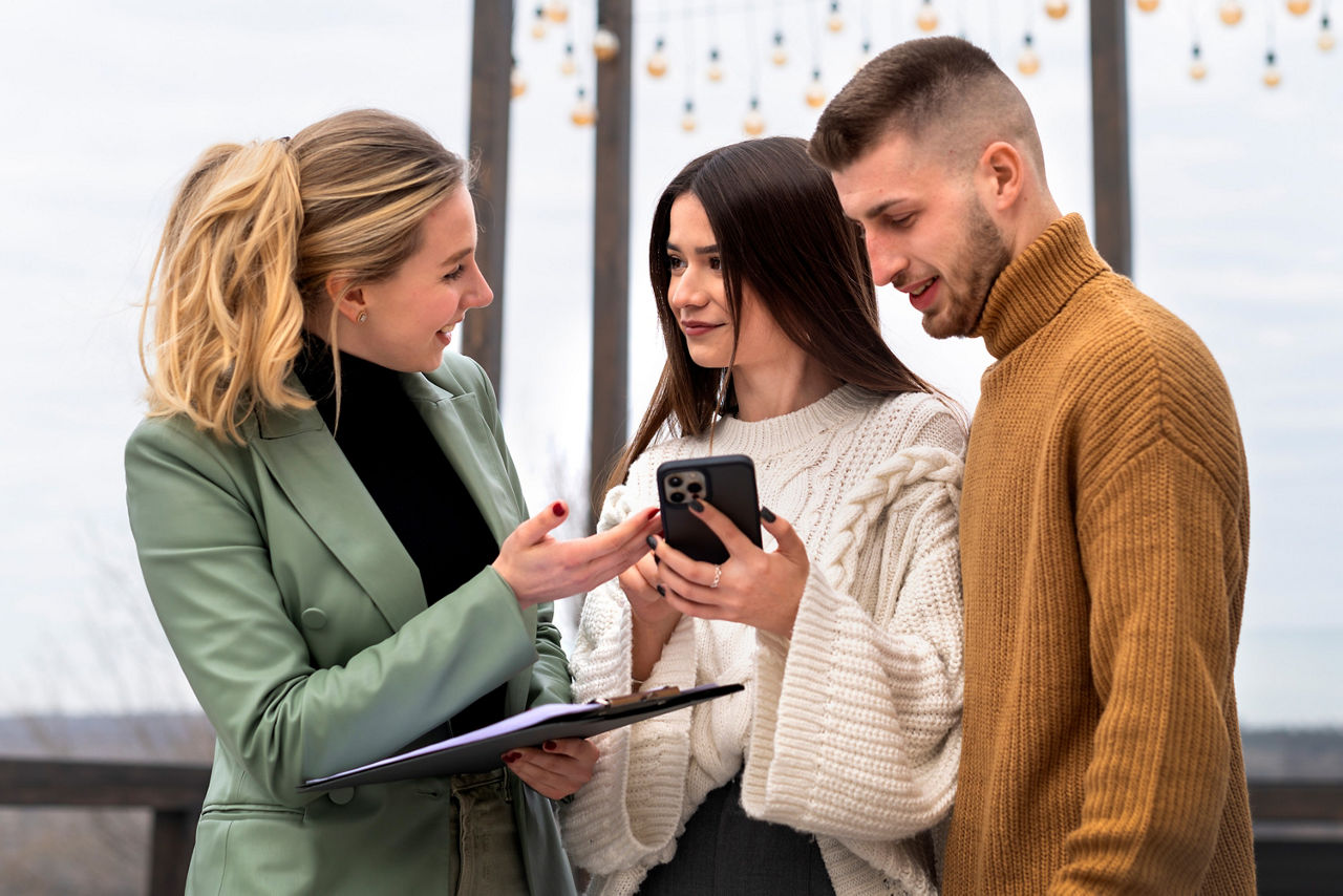 Drei junge Erwachsene im Gespräch, eine Frau zeigt auf ein Smartphone, das eine andere Frau in der Hand hält. Die Szene vermittelt den Eindruck einer Beratung zu einer digitalen Anwendung.