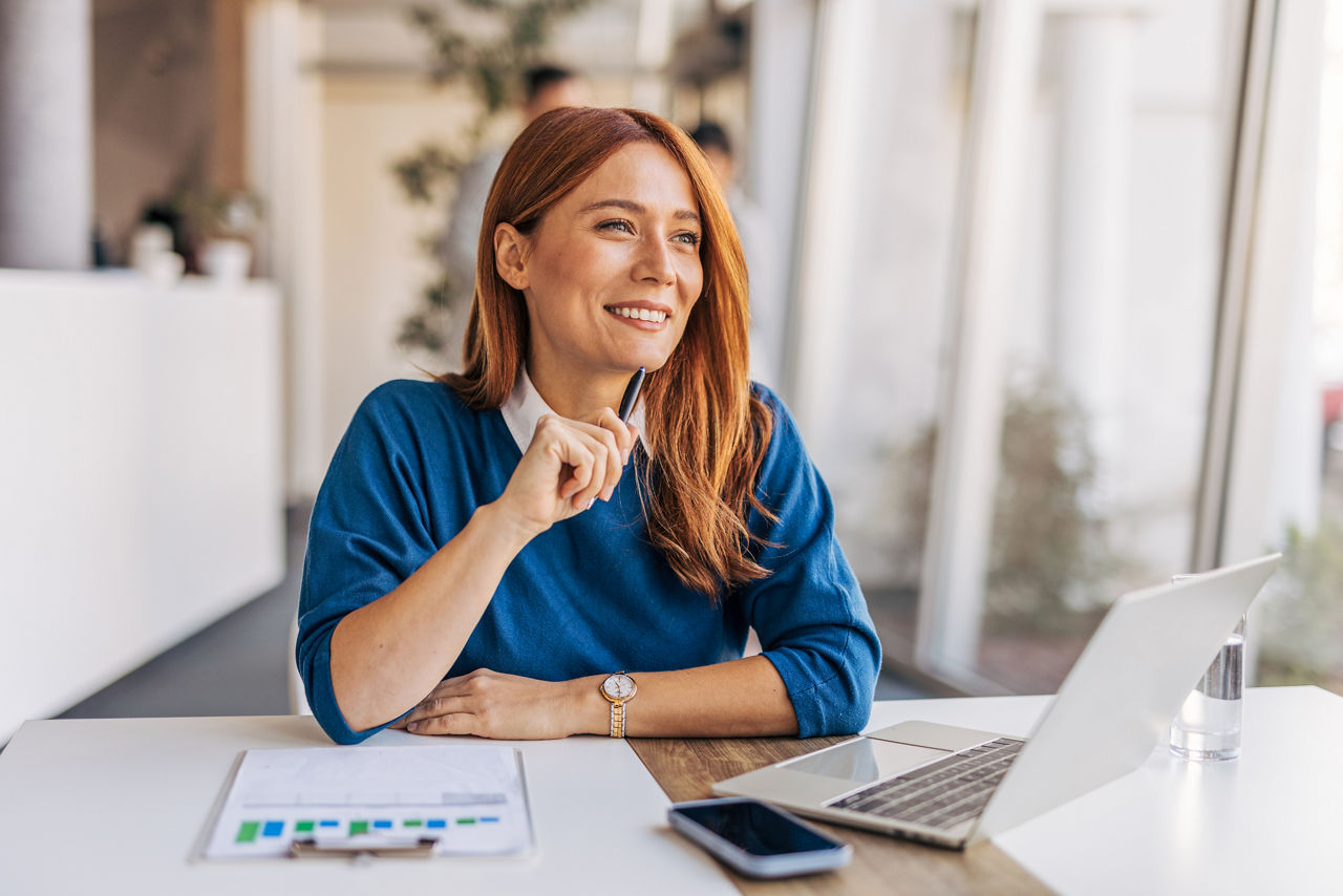 Frau sitzt mit ihrem Laptop an einem Tisch, denkt nach und lächelt dabei.