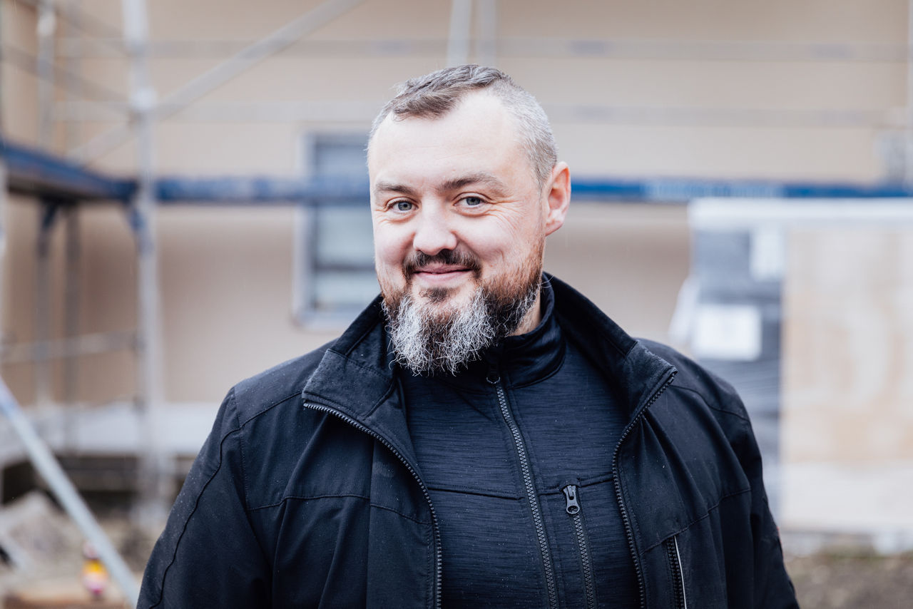 Friedrich Wilhelm befindet sich auf einer aktiven Baustelle eines Wohnbauprojekts der  „Friedrich Bau“. Er ist umgeben von Bauelementen und Gerüststrukturen, die den aktuellen Baufortschritt und das Arbeitsumfeld im Wohnungsbau widerspiegeln